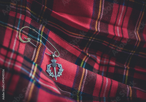 A vintage brass thistle pendant on a pin against the background of a traditional scottish kilt. Symbols of Scotland. Selective focus.