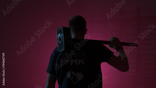Man with a sledgehammer behind his back. Guy with a sledgehammer in the red room. Man with a big hammer or a sledgehammer behind comes into the room with a red light. Muscular man with hammer from