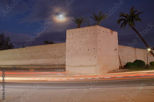 Widok nocnego Marrakeszu w Maroku, zabytkowy mur wokół medyny, palmy, poświata księżycowa, księżyc wygląda zza chmury, rozmyte światła samochodów, długie naświetlanie