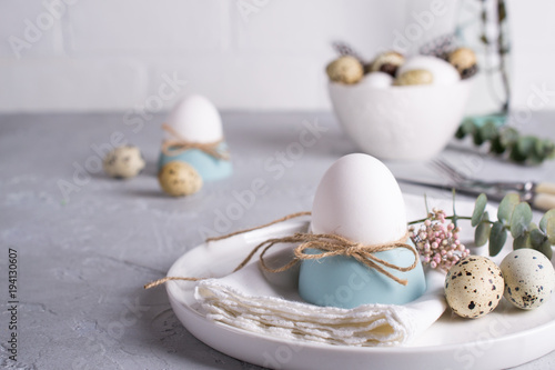 Easter festive table setting with white chicken eggs in eggs cups, leaf sprigs of eucalyptus. On a gray concrete background.