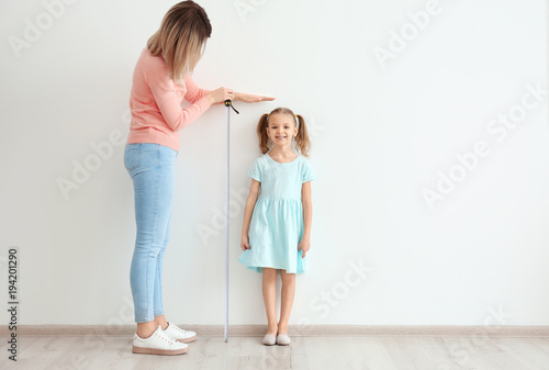 Mother measuring height of little girl near light wall