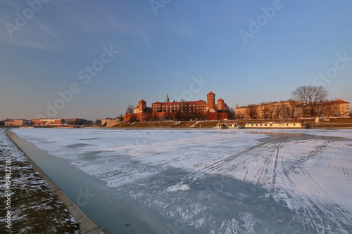 Piękny zimowy pejzaż zamku na Wawelu w Krakowie z zamarzniętą rzeką Wisłą na pierwszym planie, światło zachodzącego słońca, na lodzie na rzece ślady sanek i butów