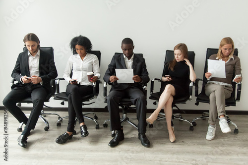 Nervous applicants preparing for employment interview sitting on chairs in queue row line, african and caucasian diverse unemployed people feeling stressed about tedious waiting, job hunting concept