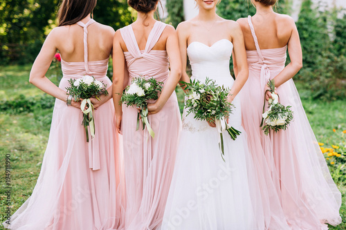 three bridesmaids in powdery dresses transformers with bouquets in hands stand with their backs near the bride in a white dress with a wedding bouquet in her hand on a green lawn