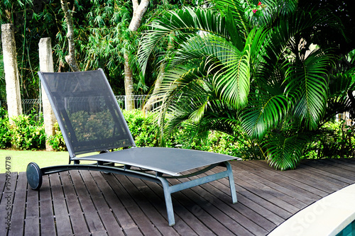 A chaise longue stands on a wooden floor near the pool. In the background are green palm leaves. Side view with copy space