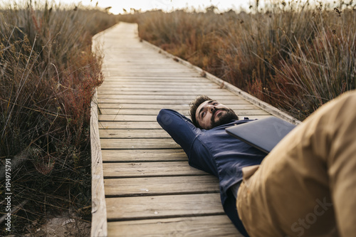 Adult man with laptop chilling on wooden pier in tall grass and enjoying freedom in sunlight.