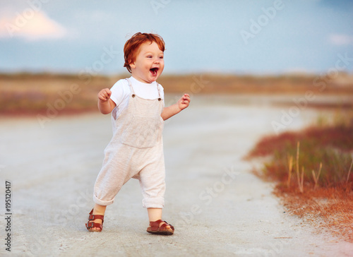 adorable redhead toddler baby boy in jumpsuit running through the summer road and field