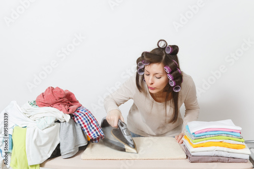 Young pretty housewife with curlers on hair in light clothes ironing family clothing on ironing board with iron. Woman isolated on white background. Housekeeping concept. Copy space for advertisement.