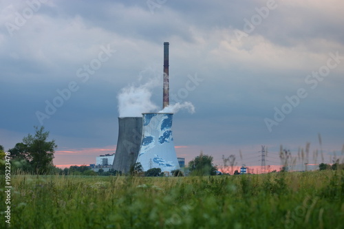 Dwa duże i jeden bardzo wysoki komin fabryczny, z dwóch wydobywa się gęsty dym, pochmurne, zasnute niebo, na pierwszym planie rozmyty rząd trawy