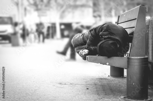 Poor homeless man or refugee sleeping on the wooden bench on the urban street in the city, social documentary concept, selective focus, black and white