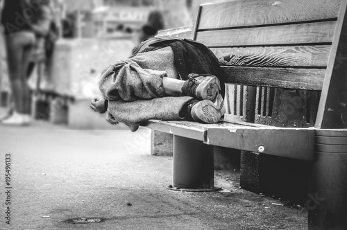 Poor homeless man or refugee sleeping on the wooden bench on the urban street in the city, social documentary concept, selective focus, black and white