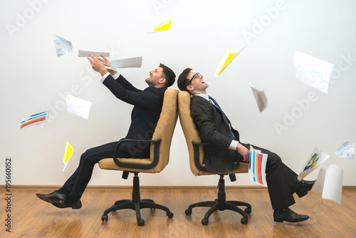 The two men sit on chairs and throwing papers on the white wall background