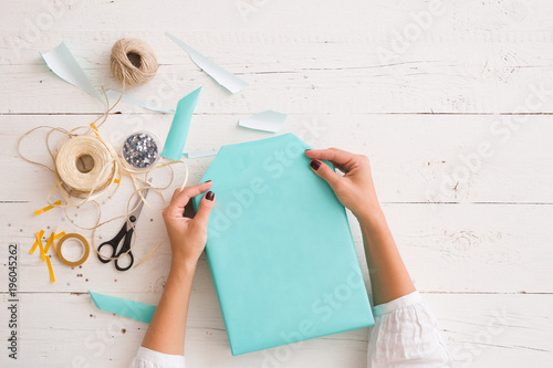 girl's hands with gift ribbon, twine, tape and present wrapped in turquoise gift paper on white wooden table background. Christmas, birthday or any other celebration preparations.