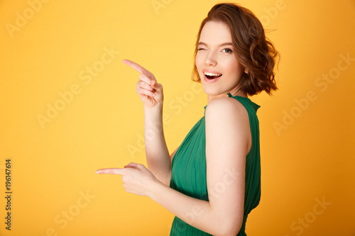 side view of attractive winking woman pointing away isolated on orange