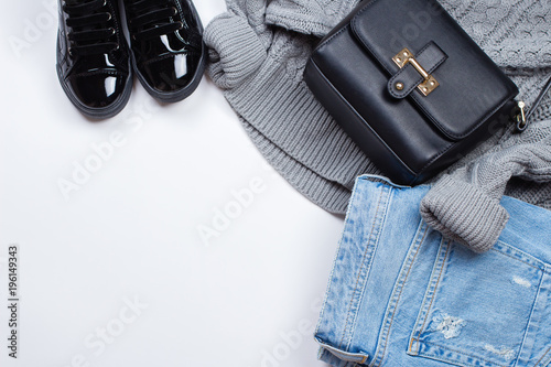 Woman Trendy Fashion Clothes. Blue jeans, knitted gray sweater, patent leather shoes boots and accessories black bag on white background, flat lay, top view, copy space