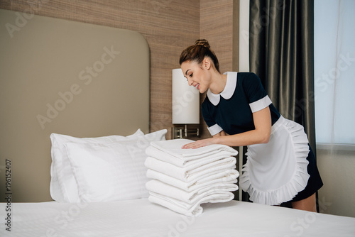 young smiling maid in uniform putting stack of clean towel on bed at hotel suit