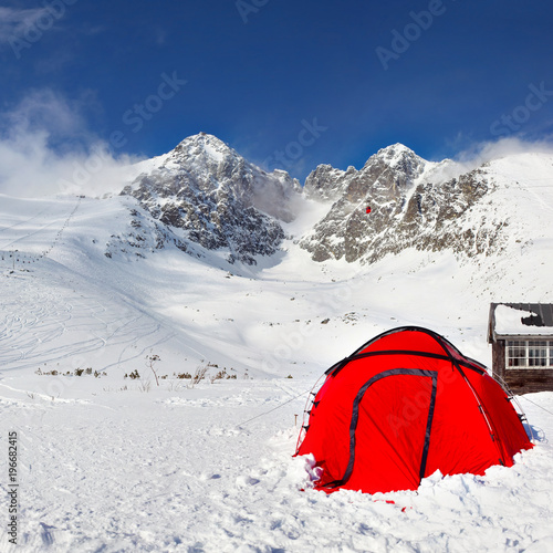 Bright red climbing tent on snow with Lomnicky Stit peak in the background, on a sunny winter day. Tatranska Lomnica ski resort, Slovakia