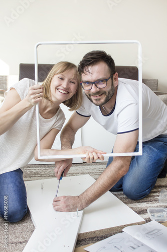 Mąż i żona skręcają meble w salonie. Uśmiechnięci pozują do zdjęcia z elementem biurka jako ramką do zdjęcia.