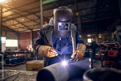 Close up portrait view of professional mask protected welder man in uniform working on the metal sculpture at the table in the industrial fabric workshop in front of few other workers.