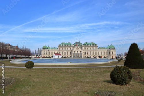 Piękny pałac w Wiedniu, stolicy Austrii, na pierwszym planie zadbany ogród z przystrzyżoną trawą i krzewami, kamienna sadzawka, błękitne niebo 