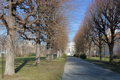 Jesienny park w słoneczny, ciepły dzień, aleja drzew, w tle widoczna historyczna zbudowa miejska, intensywnie niebieskie nebo