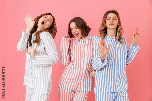 Portrait of three beautiful young girls 20s wearing colorful striped pyjamas having fun during sleepover, isolated over pink background