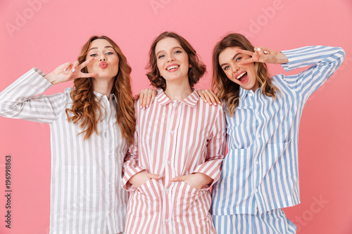 Three cheerful women 20s wearing leisure clothings having fun at slumber party and showing peace signs, isolated over pink background