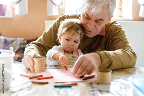 Cute little baby toddler girl and handsome senior grandfather painting with colorful pencils at home. Grandchild and man having fun together