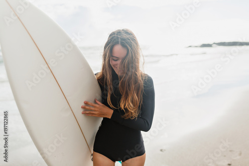 Surfer girl on the beach holding surf board watching ocean waves in sunny day. True happy emotions of young active pretty woman prepare to surfing. Summer vacation lifestyle.