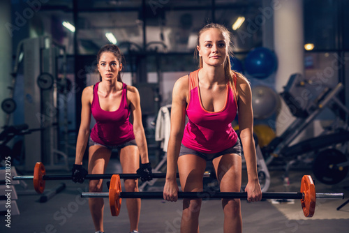 Portret z bliska widok dwóch młodych fitness zmotywowanych atrakcyjnych zdrowych sportowców aktywnych szczupłych dziewcząt kucających podczas ćwiczeń i rozgrzewki ze sztangą na siłowni.