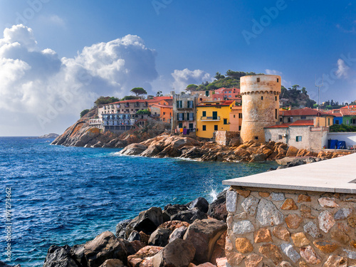 Perfect tiny seaside village of "Giglio Porto" with multi colored houses, an ancient defensive tower and a rocky coastline against a deep blue Mediterranean sea. - Giglio Island, Tuscany, Italy