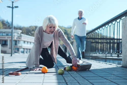 Are you ok. Selective focus on a scared senior lady standing on her knee and trying to pick up her groceries after falling down while her worried husband running to her in the background.