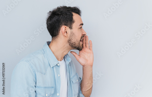 Side view portrait of attractive young Caucasian man sharing secret or whispering gossips, isolated over light grey background. Handsome male telling secret. Copy space for your advertising text.