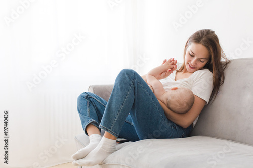 Mother is breastfeeding her kid sitting against light window background. Mom is suckling baby boy at home