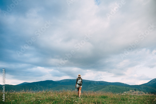 Young girl in beige hat and casual clothes with long hair is walking with cloudy sky and green mountains on background
