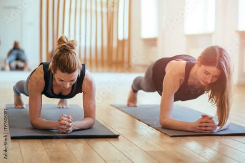 Front view of two sporty fit and slim middle aged women doing planking exercise indoors together with a natural light in modern interior sport studio hall or yoga class.