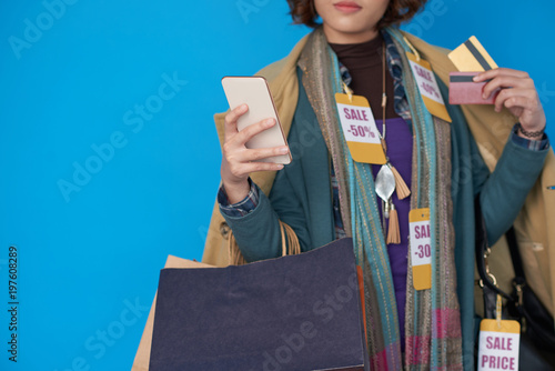 Asian shopaholic woman in clothes with sale price tags using application on her smartphone