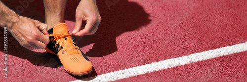 Running shoe at start line success challenge competition banner. Athlete fit man getting ready for run race tying running shoes. Panoramic on red tartan tracks.