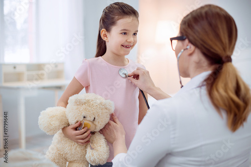 Stay for minute. Happy jolly girl holding plush bear and staying while female doctor touching stethoscope