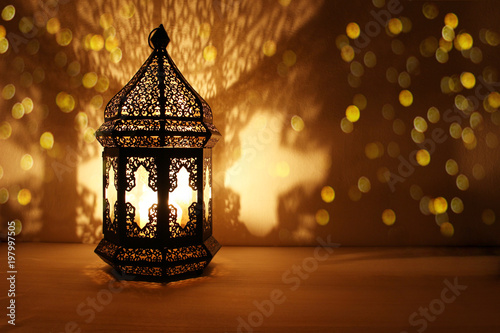 Ornamental Arabic lantern with burning candle glowing at night and glittering golden bokeh lights. Festive greeting card, invitation for Muslim holy month Ramadan Kareem. Dark background.