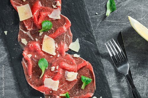 Top view of the raw beef carpaccio with parmesan cheese on a black shale service board. Delicatessen food.