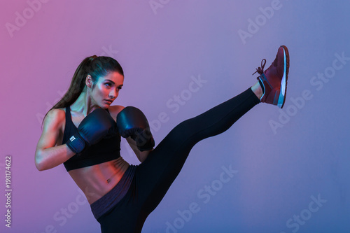 Photo of young sportswoman 20s in tracksuit and black boxing gloves fighting and kickboxing on training, isolated over dark background