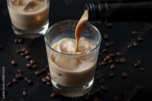 Pouring irish cream in a glass with ice, surrounded by coffee beans on a dark black background