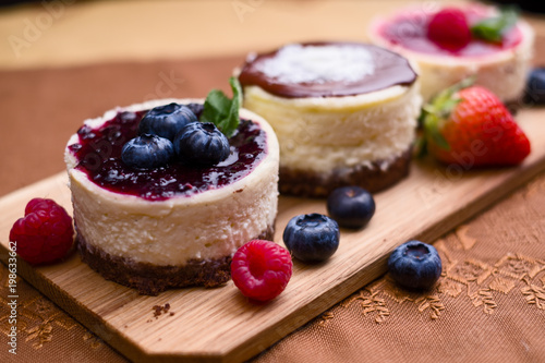 Three mini cheesecakes, one with blueberry sauce and berries decoration on brown cloth and wooden pad