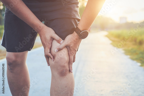 Injury from workout concept : Asian man use hands hold on his knee while running on road in the park. Shot in morning time, sunlight and warm effect with copy space for text or design