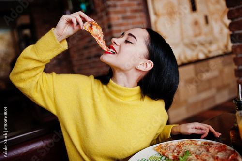 Funny brunette girl in yellow sweater eating pizza at restaurant.