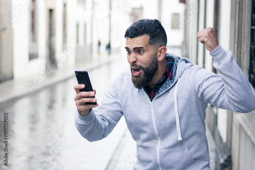 Enthusiastic young man looking at cellphone with victorious expression in the city. Overexcited hipster with grey hoodie holding mobile device and fist up. Online bet winner concept