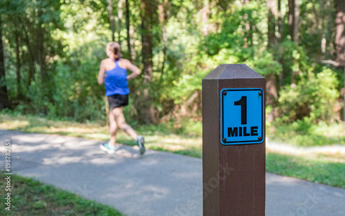 A one mile marker sign post beside a paved pathway with a bright green and sunny background and a woman running.