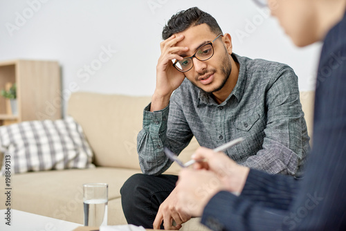 Close-up shot of young psychologist taking notes on clipboard while consulting depressed mixed-race patient during therapy session at cozy office