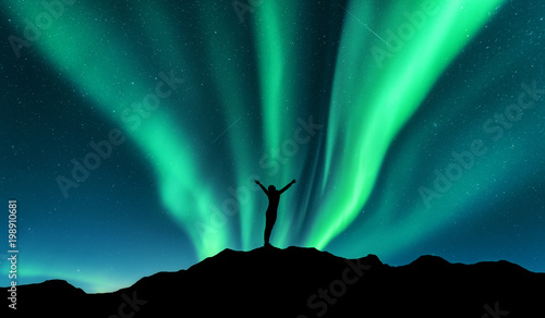 Aurora and silhouette of standing woman with raised up arms on the mountain in Norway. Aurora borealis and happy girl. Starry sky, green polar lights. Night landscape. Northern lights. Travel